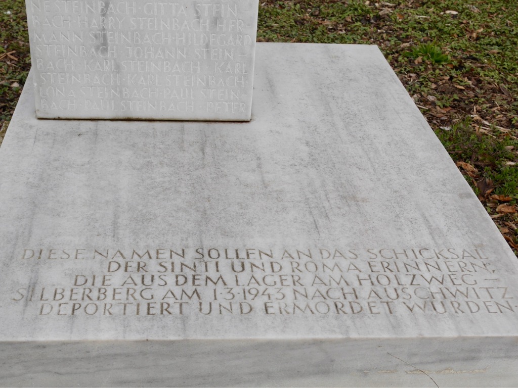 Ausschnitt aus der Namensstele für die ermordeten Sinti:zze und Rom:nja. Zu sehen ein weißer Marmorblock mit der Inschrift: "Diese Namen sollen an das Schicksal der Sinti und Roma erinnern, die aus dem Lager am Holzweg-Silberberg am 1.3.1943 nach Auschwitz deportiert und ermordet wurden." 
